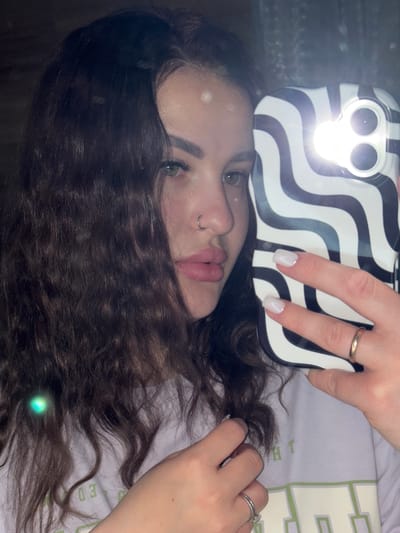 A woman lies on her back, taking a selfie with a smartphone. The phone has a black and white wavy patterned case and the flash is on. Her face is partially obscured by her hair. She has a nose ring and visible makeup. She is wearing rings on both hands. She is lying on a light purple shirt with visible writing in light green. The location is unknown, but presumed to be in Tliustenkhabl', Russia.