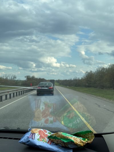 The image shows a view from inside a car on a highway. Another car is in the adjacent lane. A partially eaten bag of sunflower seeds and a bag of chips are on the dashboard. The location is likely near Berezki, Russia, given the writing on the chip bag is in Russian. The sky is cloudy.