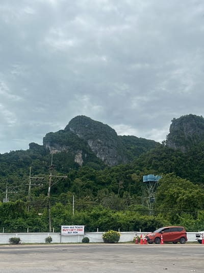 The image depicts a roadside scene in Tambon Don Sak, Thailand, featuring lush green mountains and a cloudy sky. In the foreground, an asphalt lot is visible, with a white concrete wall running along the edge of the road. A white sign attached to the wall advertises "FERRY-BUS TICKETS BUY 1 GET 1 FREE BUY NOW." To the right of the sign, a red minivan is parked, with several orange traffic cones arranged nearby. Behind the wall, dense vegetation and trees cover the lower slopes of the mountains. Power lines and poles are visible against the backdrop of the forested hills. In the middle ground, a metal water tower with three blue tanks stands amidst the greenery. The mountains themselves are characterized by steep, rocky cliffs, with trees growing on their surfaces. The overall environment suggests a coastal or island location given the ferry ticket advertisement and the presence of mountains that are common in such areas of Thailand.