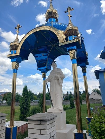 In Doslidnytske, Ukraine, a white stone statue of a man holding a book and a staff stands beneath a blue and gold canopy adorned with stars. The canopy is supported by four golden columns, each decorated with blue accents and intricate heart-shaped carvings. Above the canopy, a golden dome with multiple layers and a cross rises towards a bright blue sky filled with white, fluffy clouds. Several smaller, similarly adorned domes with crosses are visible on the sides. The statue is placed on a white pedestal. In the background, a green lawn is surrounded by trees and a grey stone fence. A building with blue and yellow elements, typical of Ukrainian architecture, is partially visible on the right. The scene appears to be a religious site, possibly part of a church or a memorial.