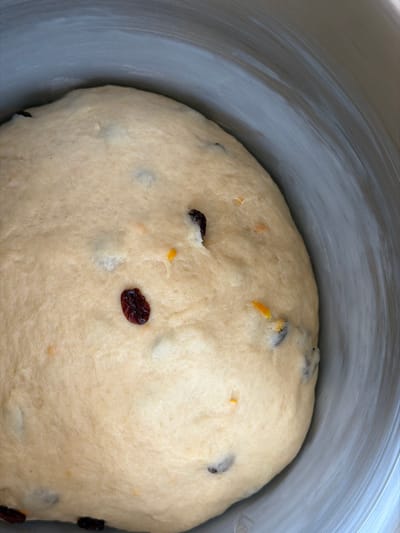 A close-up, top-down view reveals a light tan, opaque dough mass occupying the lower portion of a round, light gray-white bowl. The dough exhibits a smooth, slightly irregular surface, consistent with a rising process. Embedded within the dough are numerous dark reddish-brown, ovoid inclusions, likely dried fruit such as cranberries or raisins, along with smaller, irregular bright orange fragments, consistent with citrus zest or candied peel, and occasional small white pieces. The interior surface of the bowl above the dough displays faint, concentric or swirling marks. No individuals, explicit actions, or external environmental details beyond the bowl are visible. This scene is located in Bretton, United Kingdom.