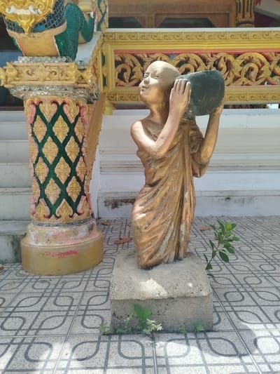 A statue of a young monk, with a shaved head and wearing golden robes, stands on a concrete pedestal in front of a decorated pillar. The monk is holding a dark bowl with both hands. The pillar is ornate, with intricate patterns in green, gold, and pink. Behind the pillar is a white wall and stairs. The ground is tiled with a geometric pattern. A small green plant is growing near the base of the statue. The scene is outdoors, under sunlight casting shadows. The statue appears to be made of a weathered material, possibly bronze or resin. The overall setting suggests a religious or cultural site in Baan Tha Po, Thailand.