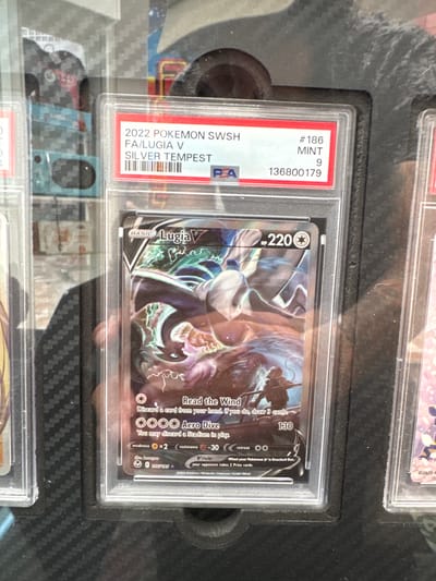 A PSA-graded Pokemon card of Lugia V from the Silver Tempest set is displayed. The card is in a clear protective case, with a red and white PSA label on top. The label indicates the card is #186, MINT, graded a 9, with the serial number 136800179. The card itself features an illustration of Lugia with stormy effects. The surrounding environment is dark, with some reflected light suggesting an indoor display case. The location of El Paso, United States of America, is not discernible from the image.