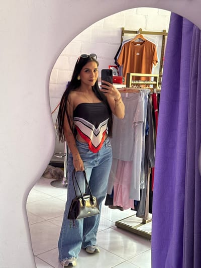 A female subject is depicted in a mirror selfie taken within a retail establishment located in Timaná, Colombia. The subject, with long dark hair and wearing dark lipstick, stands facing the mirror, holding a black smartphone with her right hand to capture the image and a black structured handbag with her left hand. She wears a strapless bandana-style top patterned in black, white, grey, and red, light blue wide-leg jeans with frayed hems, and light-colored athletic shoes. Sunglasses are positioned on her head, and she wears gold-colored stud earrings and multiple red string bracelets on her left wrist, with tattoos visible on her left forearm. The background, reflected in the mirror, displays a white textured wall on the left, with a silver-toned bar stool partially visible at the lower left. To the right, a gold-colored clothing rack system holds various garments, including an orange t-shirt with an "alo" logo. A red portable speaker rests on a shelf behind the subject's head. A purple textured curtain extends vertically along the right edge of the frame, possibly indicating a fitting room or display section. The flooring consists of white square tiles.