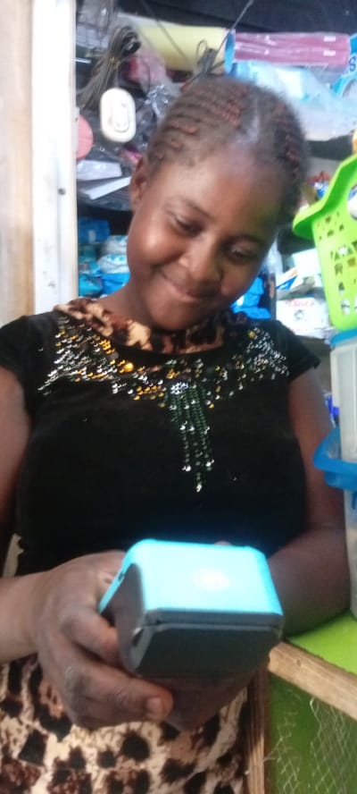 An indoor portrait captures a woman in Kwachiri, Nigeria, engaging with a small electronic device. She is smiling gently and looking down at a light blue and black portable device, which she holds in both hands. A white circular logo or icon is visible on the top surface of the blue part of the device.
The woman has dark skin and her hair is styled in tight, short braids. She wears a black short-sleeved top with a brown and black leopard print collar. The front of her top is adorned with numerous gold, silver, and green sequin-like embellishments. A portion of a brown and black patterned skirt or lower garment is visible below her hands.
The background is somewhat cluttered, suggesting a functional space, possibly a small shop or a domestic area. Various items are visible on shelves behind her, including bundled wires, what appears to be a white electrical component hanging on the left, and blue and green plastic containers. A bright green plastic basket is positioned to the right of her head. On a blue package in the background to the right, the letter "S" is faintly discernible. The overall lighting is consistent with an indoor setting.