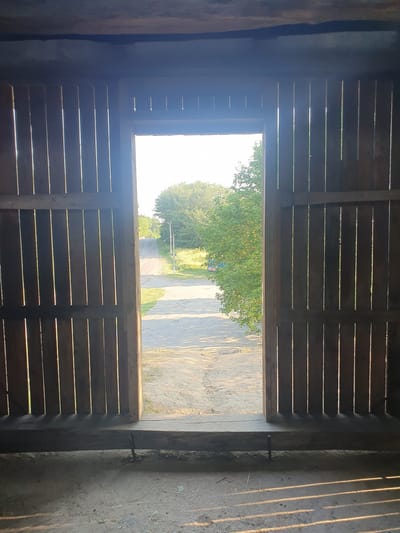This media file depicts a view from inside a rustic wooden structure, looking out through an opening. The structure appears to be made of weathered wooden planks, with horizontal slats forming the walls and ceiling of the interior, allowing slivers of sunlight to penetrate.
The opening reveals an outdoor scene bathed in the warm light of what appears to be late afternoon or early evening. Sunlight casts long shadows across a gravelly path or yard outside. Lush green trees are visible in the mid-ground, their leaves dense and full. Beyond the trees, a road or path winds its way into the distance, flanked by more greenery and what looks like a utility pole. A small, colorful object, possibly a vehicle or piece of equipment, is situated near the road, adding a touch of contrast to the natural landscape.
The overall impression is one of a rural or semi-rural setting, perhaps a barn, shed, or open-sided shelter. The time of day is suggested by the low angle of the sun and the long shadows. The weather appears to be clear and fair. The image captures a moment of quiet observation, looking out from a sheltered space into the open world. There is no visible text in the media file. The location context provided is Halych, Ukraine.