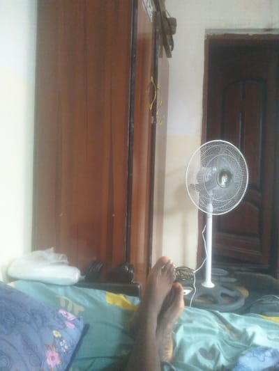 The photograph shows a room interior. A bed is in the foreground with a blue pillow and a light blue and green patterned sheet. Two human feet are visible on the bed. Behind the bed, on the left, is a brown wardrobe. On the right is a standing white electric fan, with a brown wooden door partially visible behind it. A white wall is present. The location is Gauta, Nigeria, as indicated by the query.