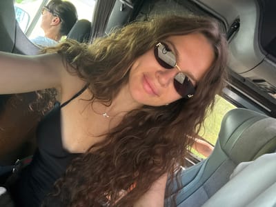 A selfie shows a young woman with long, wavy brown hair and sunglasses with gold frames, wearing a black top and a delicate necklace. She is in a vehicle, possibly a bus or van, with light gray seats visible in the foreground and background. Sunlight filters in from a window, illuminating the scene. Another person with dark hair and glasses is visible in the background, looking out the window. The location is stated to be in Onguday, Russia.