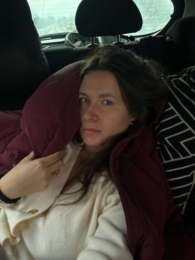 A young adult female is depicted lying or reclined in the back seat of a vehicle, reportedly in Рассудово, Russia. She has long, dark brown hair and light-colored eyes, looking directly towards the camera with a neutral expression. Her complexion is light, showing some minor skin variations. She is wearing a cream-colored, V-neck, ribbed knit cardigan, with a single gold-colored button visible on the left side of her chest. Her left hand is visible, partially holding a maroon-colored puffer garment, which appears to be a vest or jacket, and a silver-toned ring with a central clear stone is present on her left ring finger. A small gold stud earring is visible in her right ear. To her right, a black and white geometrically patterned textile, possibly a pillow or blanket, is partially visible. The dark interior of the car is visible around her, including black headrests and portions of the rear seating. Through the rear window, a blurred outdoor scene suggests a road or similar environment under overcast conditions, with distant foliage discernible.