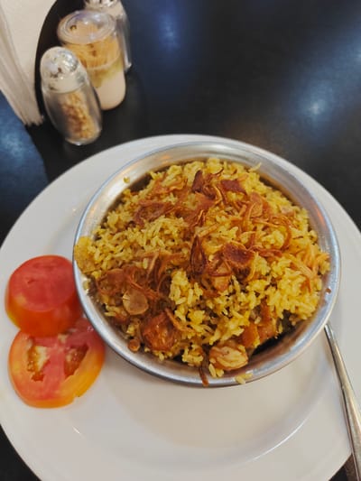 This media file is a close-up, eye-level shot of a dish of biryani served in a metallic bowl. The biryani is a vibrant yellow, garnished with crispy fried onions. Two slices of fresh tomato are placed on the side of the white plate. In the background, out of focus, are salt and pepper shakers, a napkin, and a dark, possibly wooden, table. The lighting suggests an indoor setting, likely a restaurant, with no specific time of day or weather indicated. The focus is entirely on the food, highlighting its presentation and textures. The location context is Chor Sahapur, Bangladesh.