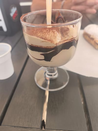 A close-up shot shows a dessert in a stemmed glass on a dark wooden table. The dessert appears to be a layered mousse or ice cream with chocolate sauce drizzled on top and dusted with cocoa powder. To the left of the dessert is a small white disposable cup. In the background, out of focus, is a person's arm and hand, suggesting they are seated at the table. The setting appears to be an outdoor cafe or restaurant in San Salvo, Italy, given the context. The overall lighting is bright, indicating it is daytime.