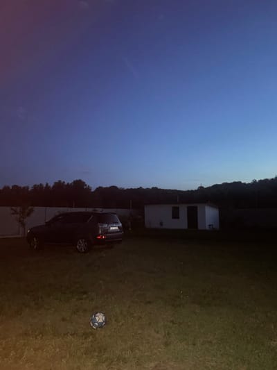 The scene depicts a twilight or early night landscape in Petrivka, Ukraine. The sky, occupying the upper two-thirds of the image, presents a deep indigo blue at the zenith, gradually transitioning to a lighter blue-grey near the horizon, with subtle hints of red-purple haze on the far left. The foreground consists of a sparsely mown grassy area, illuminated by an unseen light source, casting a yellowish-green hue on the ground. A blue and white patterned soccer ball rests on the grass in the lower center.
In the midground, a dark-colored SUV, consistent with a Mitsubishi Outlander, is parked facing right on the grassy terrain. Its rear features illuminated taillights and a visible Ukrainian license plate, reading "BH 0051 IB", indicating the Odesa region. Behind the SUV, a light-colored, possibly concrete, perimeter fence stretches horizontally across the scene. Small, dark, sparse vegetation and trees are visible both behind and in front of the fence line on the left.
To the right of the SUV and fence, a small, white, rectangular, single-story utility building or shed is visible, featuring a dark door and a rectangular window. A small, dark object, resembling a bench or stool, sits directly in front of the building's right side. Behind this structure and extending across the right side of the frame is a dense line of dark, silhouetted trees, suggesting a wooded area or forest. The overall atmosphere is devoid of human presence or activity.