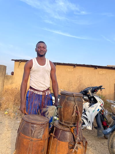 The image is a vertical portrait-style photograph, rotated 90 degrees clockwise, showing a man with traditional drums outdoors under a clear blue sky.
In the foreground, a Black man is standing, facing slightly to his left and looking directly at the camera. He has short, dark hair and a neatly trimmed beard. He is wearing a white sleeveless undershirt (tank top) and a blue garment around his waist, which appears to be a skirt or wrap, adorned with multiple brown, fringe-like strips hanging down. His arms are bent at the elbows, and his hands are positioned near the drums. In his right hand, he holds a white, fluffy, brush-like object, possibly a drumstick or a ceremonial whisk.
To the right of the man, and partially behind him, are several large, barrel-shaped drums made of wood with animal hide stretched over the ends, secured with ropes or leather straps. There appear to be at least three or four drums. One is positioned horizontally in front of the man, and others are stacked or placed nearby. The drums show signs of wear, indicating age and use.
In the mid-ground, behind the drums and to the right of the man, a white and black scooter or small motorcycle is parked. Its front wheel, handlebars, and seat are visible, along with some mechanical parts.
The background features a simple, light brown or tan building with a dark, corrugated roofline visible at the top. The wall of the building appears to be made of mud brick or stucco. Below the building, the ground is dry and dusty, with some sparse vegetation or debris visible. In the distance, between the man and the building, a small portion of another person's head and shoulders can be seen peeking out from behind a lower wall or structure.
Above the building and the man, a large expanse of clear, bright blue sky with a few wispy white clouds is visible, suggesting it is daytime with good weather. The lighting indicates it is likely midday or early afternoon, with clear sunlight. No text is visible in the image.