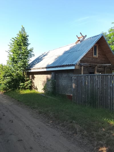 The image shows a rural scene with a wooden house and a dirt road. The house has a corrugated metal roof and is partially constructed with logs. There is a small window on the upper floor, and scaffolding or construction materials are visible on the roof. A wooden fence runs along the side of the property, and there are trees and overgrown grass. The dirt road in the foreground shows tire tracks. The sky is clear and blue, suggesting a sunny day. The location is identified as Патоцкі сельскі Савет, Belarus. There are no people visible in the image.