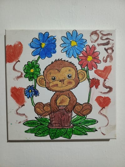 A square canvas displays a child's painting of a cartoon monkey seated in a brown wooden bucket-like object surrounded by green leaves. The monkey has brown fur, large brown eyes, and a smiling expression. It's encircled by a semi-circular arrangement of colorful flowers - blue, red, and green. Red hearts and brown squiggles are painted in the background. The name "OSCAR" is painted in brown on the upper right corner. The painting is positioned against a plain white wall. There is no evidence to establish this image's location in Quevedo, Ecuador.