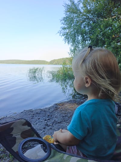 A young child with blonde hair pulled into a small ponytail sits facing right in a camouflage camping chair. The child wears a blue t-shirt and is partially visible. They are looking out at a large body of water, a lake, with calm, rippling surface reflecting the sky and trees. The shoreline is a mixture of dark sand and small stones. In the background, a dense green forest covers the rolling hills on the opposite side of the lake. The sky is a clear, pale blue. The overall scene suggests a peaceful, natural setting, likely in Nepryakhinskoye Sel'skoye Poseleniye, Russia, during daylight hours.