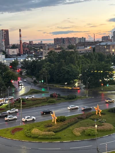 The image depicts a cityscape at dusk, likely in Tarasovka, Russia, with a focus on a wet, multi-lane intersection. The sky displays a gradient of colors from pale yellow and pink near the horizon to darker gray clouds overhead. In the background, several multi-story buildings and a prominent red and white striped smokestack are visible, suggesting an industrial or urban setting.
The foreground and middle ground are dominated by a curving road, slick with rain, reflecting the streetlights and traffic signals. Several vehicles are present on the road: a white van is turning at the intersection, illuminated by its headlights; a white sedan and a dark sedan are driving in the same lane, also with headlights on; another white sedan is in an adjacent lane. Further down the road, a silver SUV is visible. The intersection is marked with painted white lines and has traffic lights. One traffic light is red, while another is green.
The roadside is lined with trees and green landscaping. Decorative red star-shaped lights, illuminated, are positioned on the landscaped islands within the intersection and along the roadside. One star is particularly prominent in the lower right quadrant of the image. The overall impression is one of a wet, busy urban intersection during twilight, with decorative lighting adding a festive touch.