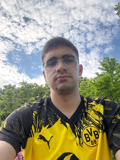 In Hattersheim, Germany, a young man wearing glasses and a Borussia Dortmund football jersey is pictured outdoors under a partly cloudy sky. The man is looking towards the camera with a neutral expression. Lush green trees form the background, with a hint of pink flowers visible in the lower left corner. The image is a selfie, taken from a slightly low angle.