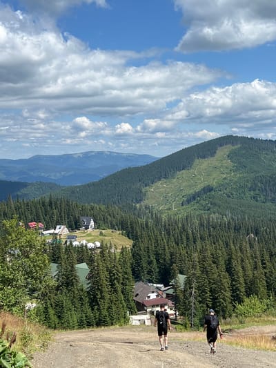 In Chorna Tysa, Ukraine, two individuals walk down a dirt path. The person in the foreground on the left wears a black t-shirt and shorts, carrying a backpack. The person on the right, also in black attire and carrying a backpack, holds a red bottle. They are surrounded by a dense forest of tall evergreen trees. In the mid-ground, several buildings with green roofs are visible, nestled among the trees. A wider mountain range stretches into the distance, with a lighter-colored slope on the right mountain indicating a ski run or cleared area. The sky is a bright blue with scattered white clouds. The overall scene depicts a mountainous, wooded landscape with a few residential structures, under a partly cloudy sky.