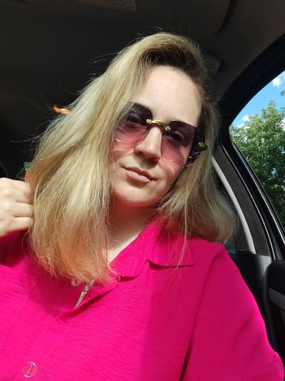 A close-up, medium shot captures a fair-skinned woman with blonde hair, wearing pink sunglasses and a bright pink collared shirt. She is seated inside a car, her left hand is partially visible, holding her hair. The background shows the car's interior and a glimpse of green trees and blue sky through the window, suggesting an outdoor, daytime setting, possibly in Vaniuki, Russia. The image is taken from a slightly elevated angle.