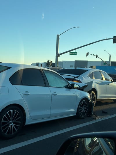 This image depicts a traffic accident scene on Metro Air Parkway in Sacramento, California. Two white cars, a hatchback in the foreground and a sedan in the background, have collided. The hatchback's front-left fender and wheel are damaged and appear to be twisted outwards. The sedan has significant damage to its rear-right quarter panel and bumper, with debris scattered on the road. Traffic lights and street signs indicating "Metro Air Pkwy" are visible, along with industrial-looking buildings and a large blue truck in the distance. The sky is clear and blue, suggesting it is daytime.