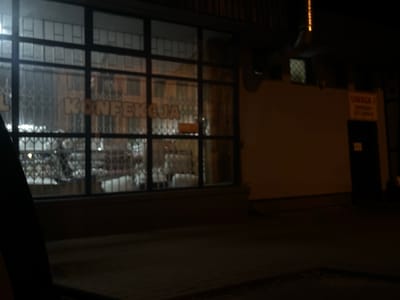 The image shows a nighttime view of a storefront with large windows in Siedlce, Poland. The word "KONFEKCJA" is visible on the window, indicating it is a confectionery shop. Inside, display shelves are visible, some with white coverings and possibly items for sale. A metal security gate is partially visible in front of the interior displays. To the right of the storefront, a wall is visible with a yellow sign that reads "UWAGA!" (Attention!) and text below it that is difficult to read but appears to be related to deliveries. A dark doorway is also present on the right side of the wall. The street in front of the store is dark and appears to be paved. The overall impression is of a closed shop at night.