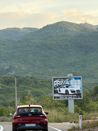 The image shows a red Hyundai i20 parked on the side of a road in Očinići, Montenegro. The car has the license plate "TV BH238". A large billboard advertising "PRIMA CAR" stands nearby. The background features a densely forested mountain and a cloudy sky. Power lines are visible stretching across the landscape. The image captures a scene along a road in a mountainous region of Montenegro, possibly an advertisement location along a scenic route.