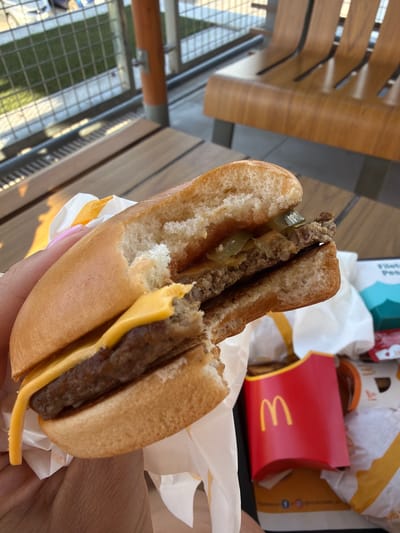 This close-up image captures a partially eaten double cheeseburger held in a hand, likely at an outdoor dining area of a fast-food restaurant in Burjassot, Spain. The burger features two meat patties, melted yellow cheese, and green pickles, with a bite already taken from its side. The hand holding the burger has a fingernail painted with light pink polish.

The meal is presented on McDonald's branded packaging, including a white paper wrapper for the burger and a red fries carton prominently displaying the golden arches "M" logo. Other fast-food packaging, such as a white tray liner and a fragment of a blue box partially revealing "Filet-O-Fish" text, is visible in the background.

The setting appears to be a modern outdoor or semi-outdoor space, characterized by light-colored wooden and dark metal slat furniture, possibly chairs or benches, and railings. In the lower left, a metal grid fence or railing overlooks a green area, suggesting a terrace or balcony setting. The bright, natural sunlight indicates it is daytime, likely a sunny day. The scene depicts a casual meal being enjoyed outdoors.