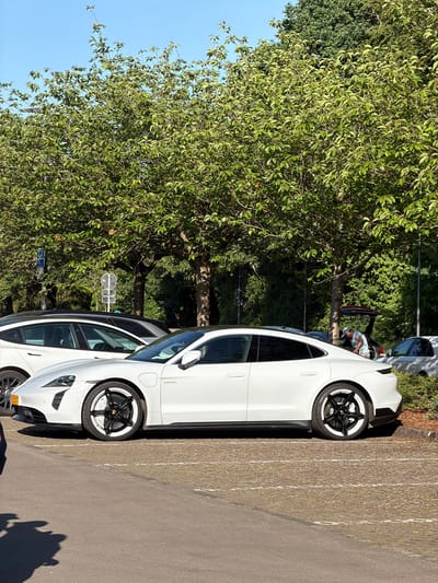 The image shows a parking area in Fentange, Luxembourg, with two white Porsche Taycan electric vehicles parked adjacent to each other. A large green tree is present on the left side of the frame, partially obscuring the sky. In the background, there are more parked cars and a person is visible near a car. The parking area is paved, and marked parking lines are present.