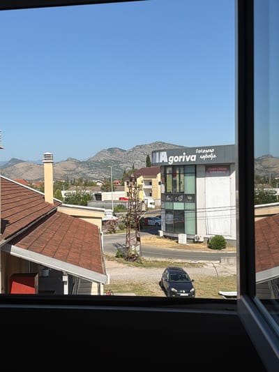 The image shows a view from a window onto a street scene in Mojanovići, Montenegro. In the foreground, the top of a dark windowsill is visible. To the right, a portion of a window frame, which is dark with a gray metallic reflection, is visible. The main scene unfolds beyond the window.

On the left side of the image, a reddish-brown tiled roof of a building is prominent. A tall, light-colored chimney with multiple horizontal vents extends upwards from this roof. Adjacent to the chimney, another building with a light-colored facade is partially visible.

Dominating the middle ground is a modern, multi-story building with a gray facade and large glass windows. A prominent sign on this building reads "IA goriva najbolja." Below this, there are several circular icons and some text that is difficult to decipher. A large white banner with some red text and images is attached to the lower part of this building. An air conditioning unit is mounted on the exterior wall of this building.

To the right of the modern building, a dark-colored sedan is parked on a grassy area. The car has its headlights on, and a license plate is visible, though unreadable. In the background, a gently sloping mountain range with scattered trees and vegetation stretches across the horizon under a clear blue sky. Several smaller buildings, some with red roofs, are scattered throughout the landscape between the foreground and the mountains. A tall, rusted metal structure, possibly an old utility pole or tower, stands near the center of the image. A street curves through the scene, with a white car visible on it in the distance. A bicycle with red accents is parked near the edge of the grass in the lower part of the image. The overall impression is a sunny day in a semi-urban or suburban area with a backdrop of natural hills.