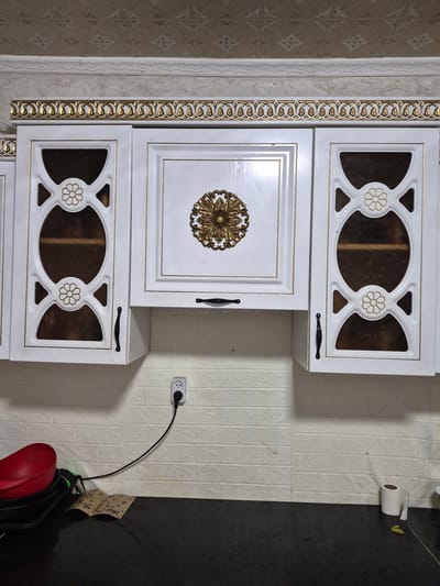 The image depicts the interior of a kitchen, likely in a residential setting. The focus is on a section of white kitchen cabinets with ornate gold trim and decorative elements on the doors. One cabinet door features a raised gold filigree design, while two others have decorative cutouts revealing tinted glass inserts, each with a gold floral motif. Black handles are visible on the cabinet doors. Above the cabinets, a patterned wallpaper and decorative trim are present. Below the cabinets, a white brick-patterned backsplash lines the wall. A black power cord runs from a white electrical outlet to an unseen appliance. On the black countertop beneath the cabinets, there are various objects including a red bowl, what appears to be a fly trap with dead insects, a roll of toilet paper, and a green item. The overall scene suggests a domestic kitchen environment with decorative elements. The location in Қанабек, Kazakhstan is not discernible from the visual information provided in the image.
