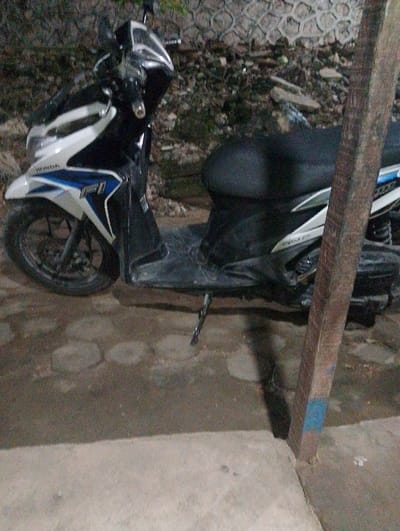 A white and black Honda motorcycle is parked on a concrete surface. The motorcycle is parked next to a large wooden post. The motorcycle is facing the right side of the image. The surface is uneven and contains several gray stones. The motorcycle is parked in Bandar Lampung, Indonesia (latitude: -5.45, longitude: 105.313). The motorcycle is in a residential area.
