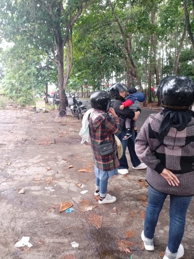 The image depicts a scene on a paved road in Kecamatan Caringin, Indonesia (latitude: -7.523, longitude: 107.485). Three people are standing on the side of the road. Two of them are wearing motorcycle helmets. One person is holding a small child in their arms. Two motorcycles are parked on the side of the road in the background. There are leaves and debris on the road. The surrounding environment is a wooded area. The sky is overcast. The scene appears to be taking place on a weekday.