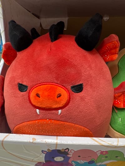 A close-up, head-on shot focuses on a red, dragon-like plush toy. The toy has two black, pointed horns on its head and two small, red, wing-like appendages with white speckles on either side of its head. Its face features black, angry-looking eyes, a textured orange snout with two nostrils, and two visible white fangs. The area around its mouth is a glittery orange. The plush toy is positioned inside a box, with the edge of the box visible at the bottom of the frame. The background is out of focus, suggesting it might be a retail environment. No specific location in Keila, Estonia, can be identified from the image.