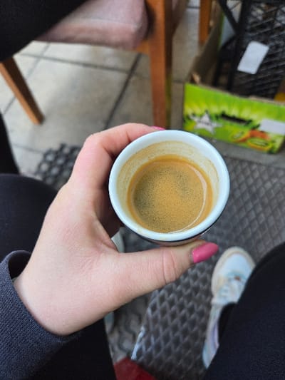An overhead, first-person perspective image captures an individual's left hand holding a small, white ceramic espresso cup filled with a shot of coffee topped with a golden-brown crema. The individual's thumb, sporting bright pink nail polish, is visible gripping the cup. The person is dressed in dark, long-sleeved attire and dark pants. A white sneaker with light blue detailing is partially visible on the bottom right, indicating the individual's foot. The ground beneath features a dark, patterned mat covering a light-colored tiled floor. In the blurred background, the leg of a wooden chair and a green and white cardboard box, possibly a produce container, are discernible. The scene is situated in Gevgelija, North Macedonia; however, the exact establishment or precise location within the city cannot be identified from the visual content.