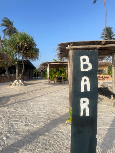 The image displays an inviting outdoor bar area located on a sunny beach in Kiwengwa, Tanzania. In the foreground, a prominent dark wooden sign with "BAR" painted in large white capital letters hangs from a rustic structure made of wooden poles and a thatched roof. The ground beneath is composed of fine, light-colored sand, showing various footprints and tire tracks.
Beyond the main sign, several other open-air structures with thatched roofs are visible, nestled amidst lush tropical vegetation, including tall palm trees and dense green plants. One tall, slender palm tree arches into the clear blue sky on the left side of the frame. Long, distinct shadows stretch across the sand, indicating either morning or late afternoon sunlight. In the background, partially obscured, another sign can be seen, which reads "Beach Lounge Zanzibar T - T".
The scene is tranquil, with no people visible, suggesting a peaceful moment at this beachside establishment. The presence of tables and chairs within the shaded structures implies an area set up for relaxation, dining, or enjoying drinks. The overall atmosphere is one of serene tropical leisure under a bright, clear sky.