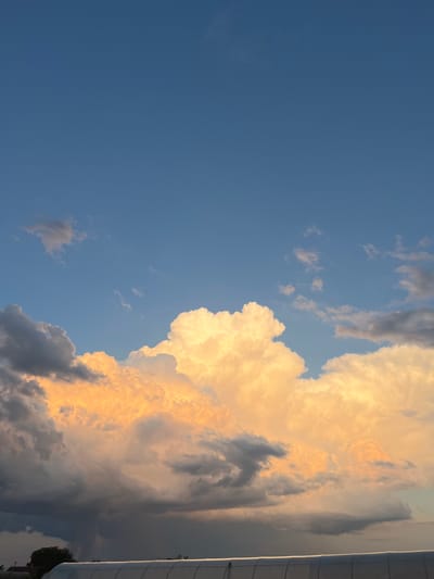 The image depicts a dramatic sky filled with cumulus clouds illuminated by the warm glow of sunset or sunrise. The clouds range in color from brilliant white and golden yellow in the foreground to darker grays and blues towards the bottom, suggesting varying depths and atmospheric conditions. A hint of a structure, possibly a greenhouse or industrial building, with a white arched roof, is visible at the very bottom edge of the frame, along with a dark silhouette of trees on the left. The sky is a clear, deep blue above the clouds, indicating good weather in the upper atmosphere. The setting appears to be an open area, possibly on the outskirts of a city, given the presence of the man-made structure and distant trees. The location is identified as Slobozia, Moldova. There are no people or specific actions visible in the image, rendering it a natural landscape scene with a focus on the atmospheric phenomena.