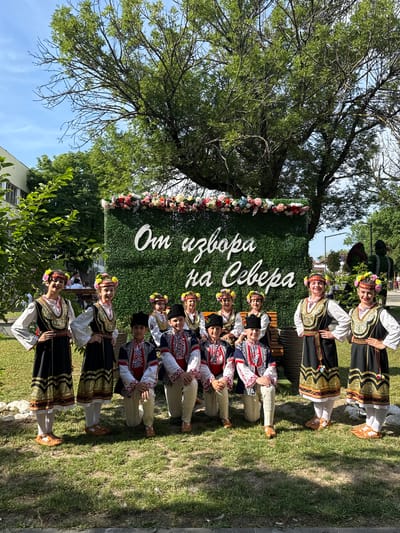 In the city of Pordim, Bulgaria, a group of ten children, all wearing traditional Bulgarian folk costumes, are posing for a photograph in an outdoor setting. The costumes consist of black dresses with gold embroidery for the girls and dark blue and red vests over white shirts and light-colored trousers for the boys. They are adorned with floral headbands. The children are arranged in two rows, with the front row kneeling or sitting on a wooden bench, and the back row standing. They are positioned in front of a large, artificial hedge-like backdrop decorated with a floral garland at the top and white lettering that reads "От извора на Севера" (From the source of the North). Behind the backdrop, a large tree with lush green leaves provides shade, and the sky is a clear blue. The ground is covered with green grass, and there are hints of other foliage and structures in the background, suggesting a park or public square. The children appear to be smiling and looking towards the camera.