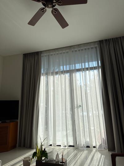The image shows an interior scene, likely a living room, in Cam Lâm, Vietnam. Sunlight streams through a large window covered by sheer white curtains and thicker grey drapes. The light casts distinct linear shadows across the floor, indicating the presence of the window blinds or the curtains themselves. A ceiling fan with dark brown blades is visible at the top of the frame. In the foreground, a small potted plant with variegated leaves sits on a surface, next to a tissue box and a white mug. To the left, a dark television screen is partially visible above a wooden cabinet with slatted doors. The overall impression is one of a bright, sunlit room with a minimalist aesthetic.