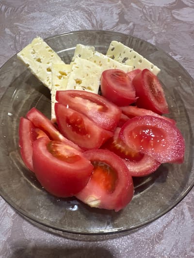 A top-down shot captures a clear glass plate filled with sliced tomatoes and cheese. The tomatoes are ripe, a vibrant red, and cut into wedges, some showing their juicy interiors. They are piled on top of thin, rectangular slices of yellow cheese. The cheese has small, irregular holes, characteristic of some varieties. The plate sits on a patterned tablecloth, its design a mix of light and darker shades, with a subtle sheen. The lighting is direct, creating highlights on the tomatoes and the rim of the plate. The overall scene is simple and domestic, suggesting a meal or snack. The image is taken in Osinniki, Russia.