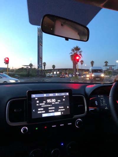 A driver is at a red light, as seen through the windshield and rearview mirror. The car radio displays FM 95.90, with the station name "KAYASS". The screen shows additional station names such as "METRO," "DING KON," and "FIVE-FM." The car's dashboard is visible with illuminated buttons and a digital display. The view outside includes palm trees, other vehicles, and buildings under a clear sky.