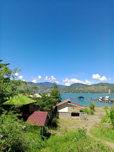 This is a landscape photograph of a body of water with floating structures, taken on a clear day. The foreground features lush green vegetation and several small buildings with red and green roofs. A light-colored building with a red-tiled roof is partially visible on the right side, with a concrete structure in front of it. Further back, a vibrant blue lake stretches out, dotted with various floating structures that appear to be houses or platforms. Some of these structures have dark blue or white roofs. The lake is surrounded by rolling hills covered in dense green trees and foliage, and in the distance, there are mountains under a bright blue sky with scattered white clouds. The scene suggests a tranquil, possibly rural, environment. The image is captured from a slightly elevated vantage point, looking out towards the lake and mountains.