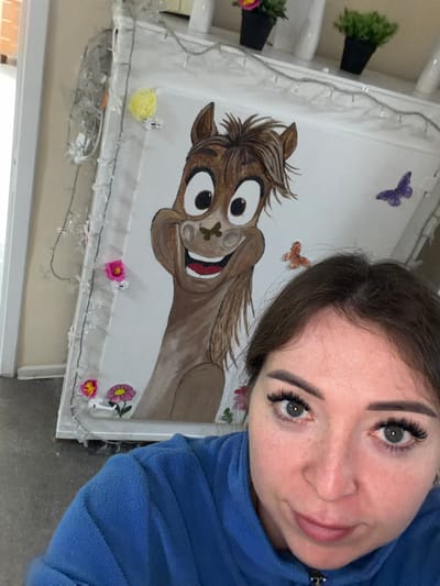 This is a medium shot, horizontally oriented selfie taken indoors. The primary subject is a young woman with dark hair, light skin, and prominent false eyelashes. She is wearing a blue fleece top and is smiling faintly at the camera. She is positioned on the right side of the frame, with her head tilted.

On the left side of the image, a large rectangular surface, possibly a wall or a large panel, is visible, adorned with a painting of a cartoonish horse. The horse has large, round eyes, a wide open mouth showing red and white, and brown fur. It is depicted in a dynamic, almost falling pose. The painting is decorated with several small, colorful butterflies and flowers, some of which appear to be three-dimensional embellishments. A string of small, white fairy lights is draped across the bottom edge of this surface.

In the background, to the left of the painting, two black planters with green artificial plants are mounted on a white surface, suggesting an indoor or sheltered environment. The overall setting appears to be domestic or recreational. The lighting is bright, indicating it's likely daytime. The woman's expression is relaxed and happy. No visible text is present in the image. The metadata indicates the location is Япрыково, Russia.