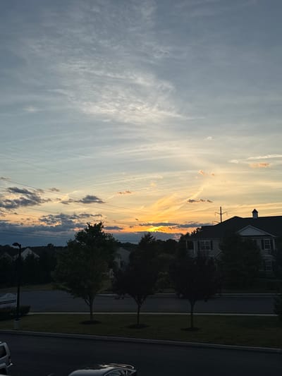 The image depicts a suburban street scene in Allen Township, USA, at dusk. A dramatic sky dominates the upper portion of the frame, with a gradient of colors ranging from pale blue and grey near the zenith to warm oranges and yellows as it approaches the horizon. Wispy cirrus clouds are visible, with some darker, more textured clouds scattered lower down. Thin power lines traverse the sky horizontally.

Below the sky, the silhouettes of houses and trees are visible against the fading light. The houses appear to be typical suburban residences with pitched roofs. Several trees, varying in size and density of foliage, are scattered throughout the scene, with some casting shadows onto the manicured lawns and paved surfaces. A street lamp is visible on the left side of the image.

In the foreground, a paved area, likely a road or driveway, is visible, with the dark shapes of at least two vehicles parked on it. One appears to be a silver SUV on the left, and a darker sedan is closer to the bottom center. The overall impression is one of a quiet evening in a residential neighborhood as daylight recedes. The image is a vertical composition, emphasizing the expanse of the sky.
