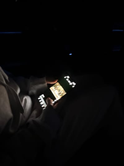 A person is holding a phone in a dark environment, possibly a car. The phone screen is illuminated, displaying what appears to be a video or image. The person's hands are visible, and they seem to be interacting with the phone. The environment is mostly dark, with some faint shapes suggesting the presence of a seat and possibly some interior features of a vehicle. The location is not identifiable as Kuchak, Armenia from the given image.