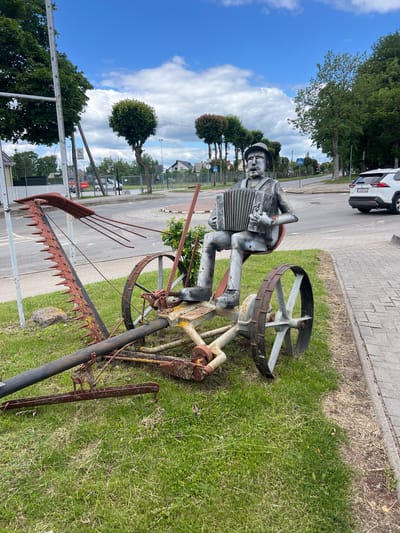 A metal sculpture of a man playing an accordion is positioned on a rusted, antique farm implement, possibly a mower, outdoors in Jurbarkas, Lithuania. The sculpture is metallic gray and depicts a man in a cap and button-up shirt, seated with his legs crossed, holding and playing a silver accordion. The farm implement itself is weathered, with visible rust and peeling paint in red and yellow. It features large spoked wheels and a long cutting bar with sharp teeth. The scene is set on a grassy area next to a paved sidewalk and a road. In the background, a white SUV is visible driving on the road, and trees and buildings line the street. The sky is partly cloudy and blue. The sculpture appears to be a decorative roadside attraction or public art installation.