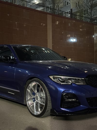 The image shows a close-up of the front of a dark blue BMW car. The car's headlight, front grille, and front wheel are visible. The wheel has a silver rim and blue accents. The car is parked next to a building with a tiled exterior. Trees are visible in the background across the street and the buildings reflect in the car's windows. The environment appears urban, suggesting the presence of streetlights and other forms of artificial illumination. The location is Novoaleksandrovka, Russia. No people are visible in the image.