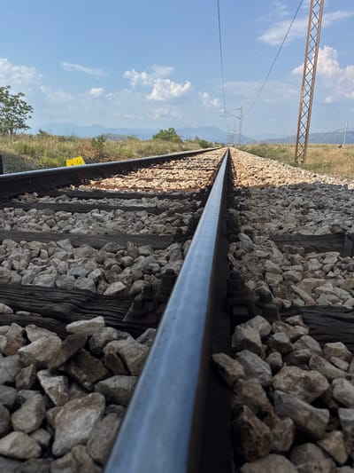 This is a low-angle, close-up shot of a railway track, taken outdoors. The perspective is from the side of the tracks, looking down the line. The foreground is dominated by the gleaming metal of the rails and the rough texture of the ballast stones that support them. Wooden sleepers are visible beneath the stones, showing signs of age and wear.

In the background, the railway line stretches into the distance, disappearing into a hazy landscape. To the left of the tracks, a bright blue sky with scattered white clouds dominates the scene. A few trees and scrubby vegetation line the side of the tracks, with rolling hills or mountains visible in the far distance. A tall, metal utility pole with crisscrossing supports is also visible in the left portion of the frame, with power lines stretching across the sky. A yellow sign with some text is partially visible on the right side of the tracks, near the middle ground.

The lighting suggests it is daytime, likely mid-morning or afternoon, with the sun casting distinct shadows. The weather appears to be clear and sunny. The overall mood is one of quiet solitude and the vastness of the landscape. The specific location is Mojanovići, Montenegro, as indicated by the provided context. No people are visible in the image, and the activity is static, focusing on the railway infrastructure itself within its natural surroundings.