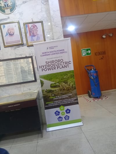 The image depicts the interior of a building, likely a government or corporate office, in Shiroro, Nigeria. The focal point is a large banner on a retractable stand, announcing "NORTH SOUTH POWER COMPANY LIMITED (NS-PCL)" and welcoming guests to the "SHIRORO HYDROELECTRIC POWER PLANT." The banner features an image of the power plant, its vision, mission, and core values.
On the wall to the left, there are framed portraits of two individuals, possibly government officials. To the right, a blue fire extinguisher on wheels stands against a wooden wall, next to a "NO SMOKING" sign and an emergency exit sign. In the foreground, a dark wooden cabinet with a metal trim is visible, with text that appears to indicate "SHIRORO PROPERTY." The floor is tiled, with visible grout lines. The overall environment suggests a formal setting, possibly an entrance or reception area, within the Shiroro Hydroelectric Power Plant or a related administrative office.
