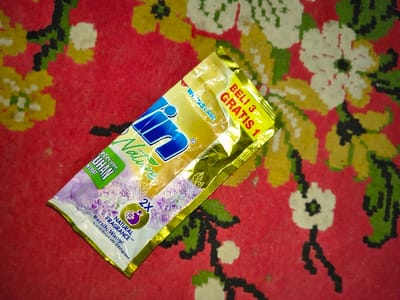 A single, intact metallic-foil pouch of "RIN Nature" detergent is resting diagonally on a red carpet. The pouch, predominantly gold with blue, green, and purple accents, displays the brand name "RIN" in blue text and "Nature" in green cursive. A yellow strip on the top right states "BELI 3 GRATIS 1" (Buy 3 Get 1 Free). Other text includes "DETERGEN KONSENTRAT," "NATURAL FRAGRANCE," and "Bersih, Wangi Lembut di Tangan," alongside an image of purple flowers and a "2X" icon. The carpet features a repeating floral pattern with yellow, white, and green elements against the red background. No individuals are present. The scene is located in Tarqumiyah, Palestinian Territory.
